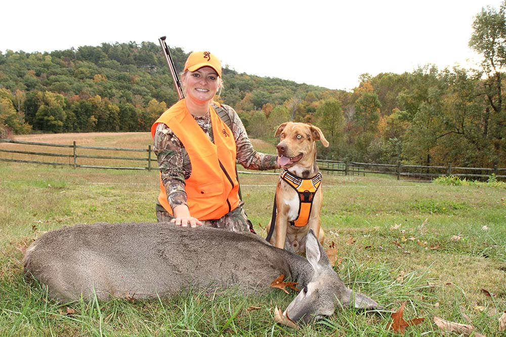 female hunter kneeling behind a whitetail doe and a hunting dog