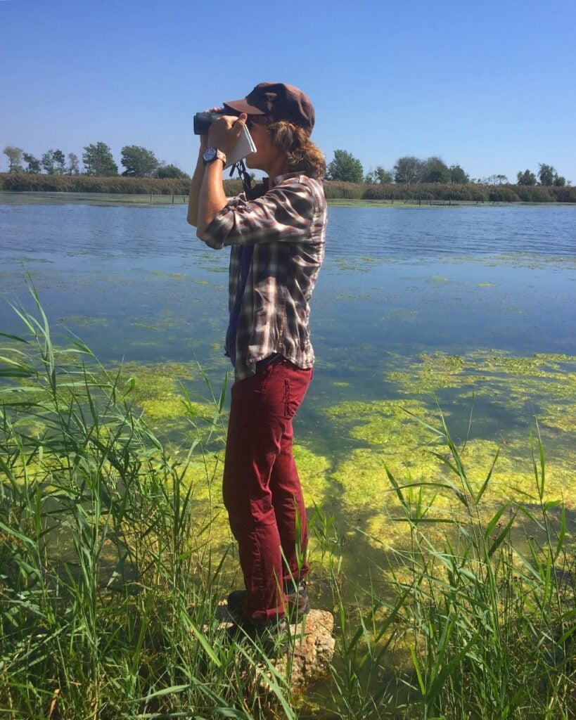 a person standing by a body of water using binoculars to look for birds