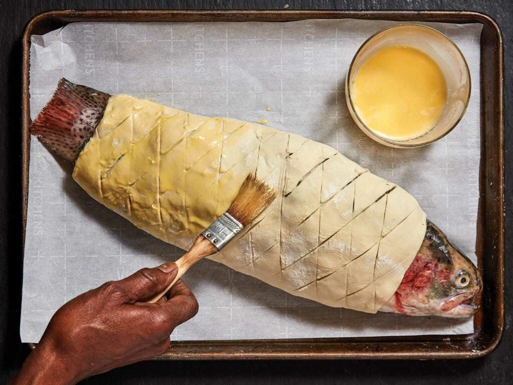 A steelhead trout wrapped in pastry dough being brushed with butter.