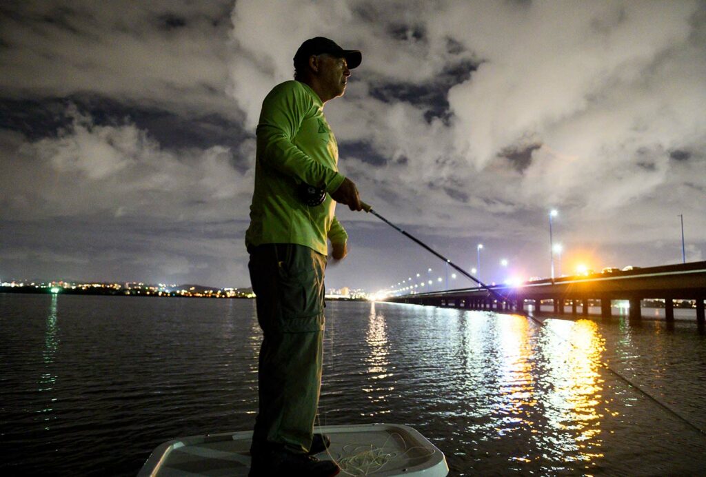 An angler fishing at sunset near a bridge.