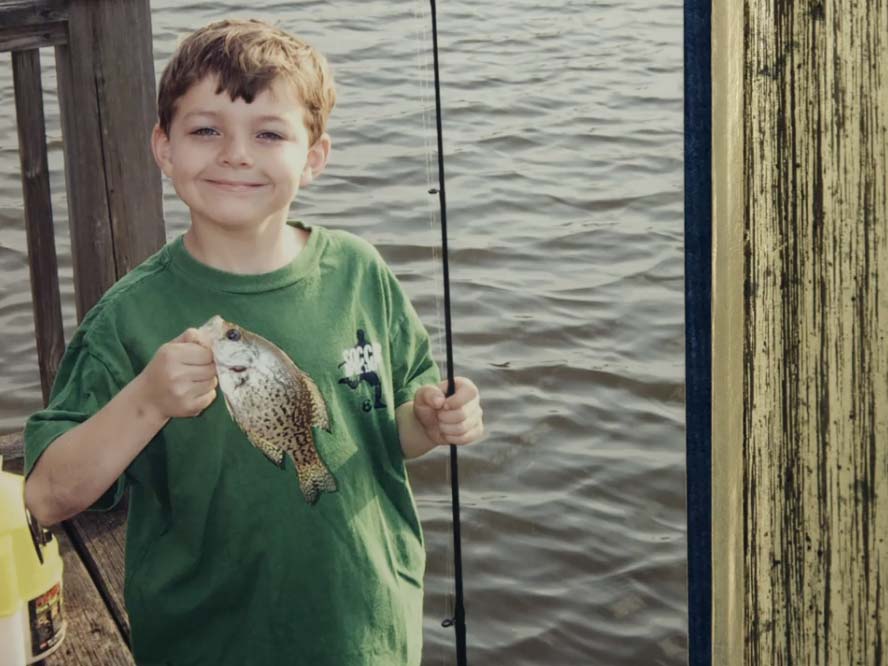 A young boy holds a fishing pole in one hand and a crappie fish in the other.