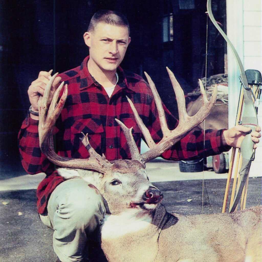A hunter in red and black flannel kneels behind the head of a whitetail deer.