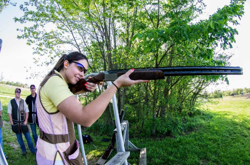 A young woman in a pink vest aims a shotgun at a shooting range.