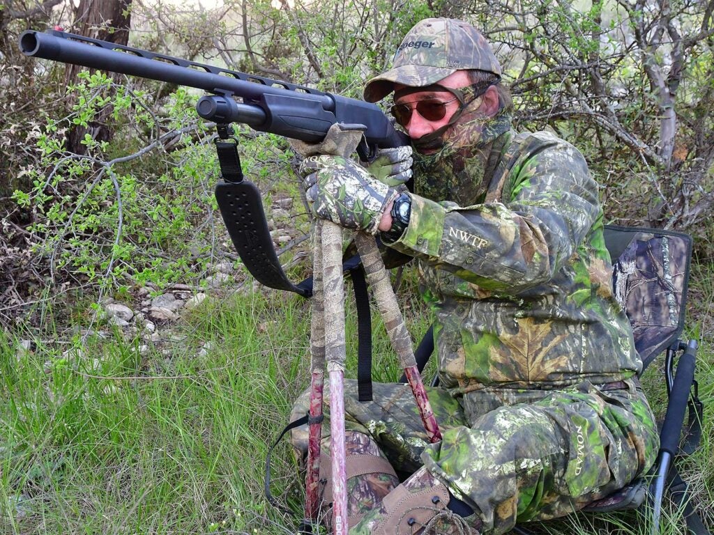 A hunter kneeling in the woods while aiming a shotgun.