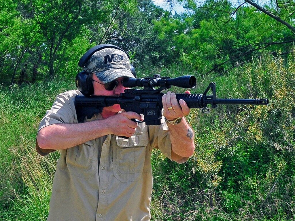A man aims an AR rifle in the woods.