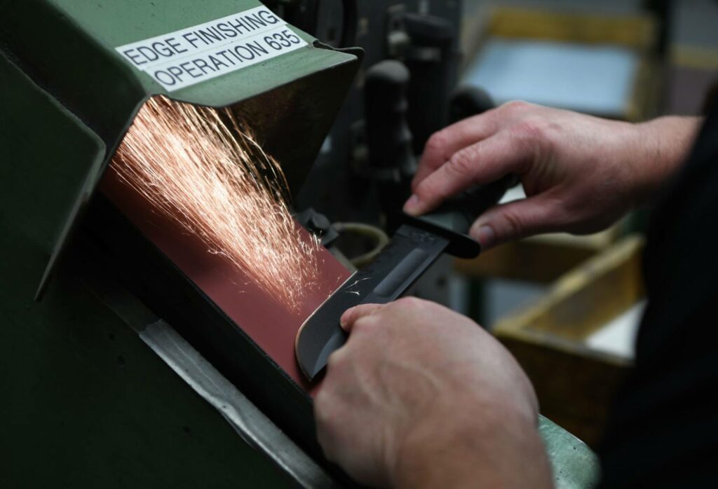 A knife maker grinds a blade of a knife on a grinding wheel.