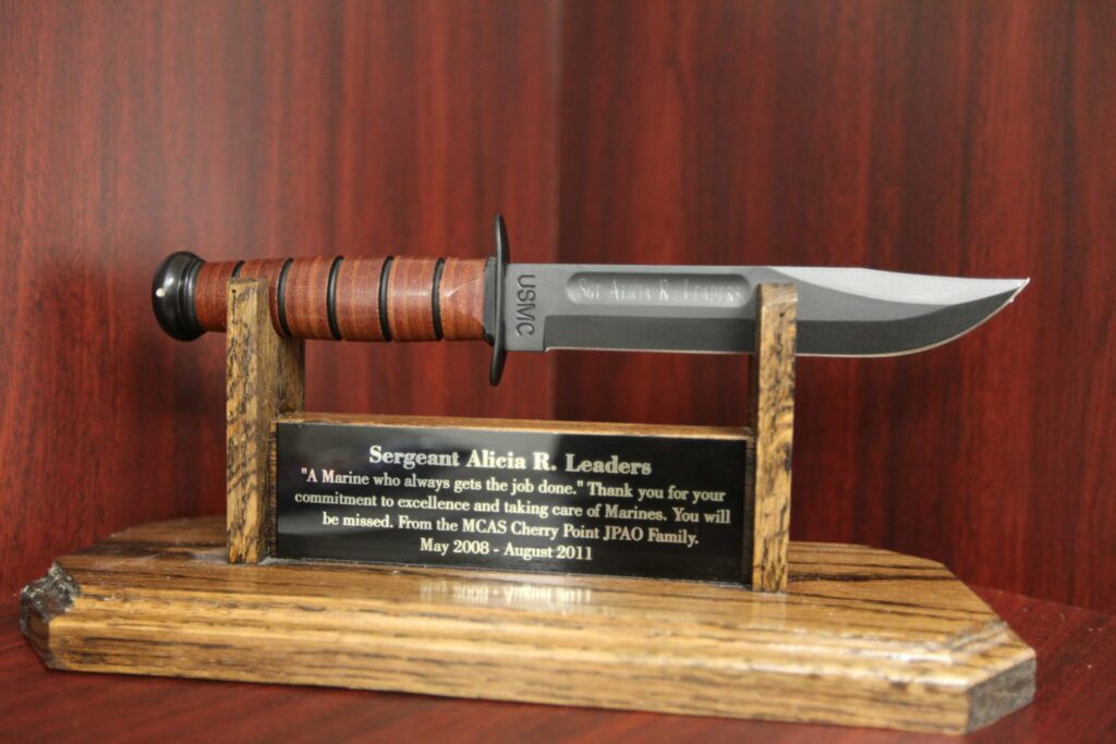 A Kabar fighting knife on a wooden stand and plaque.