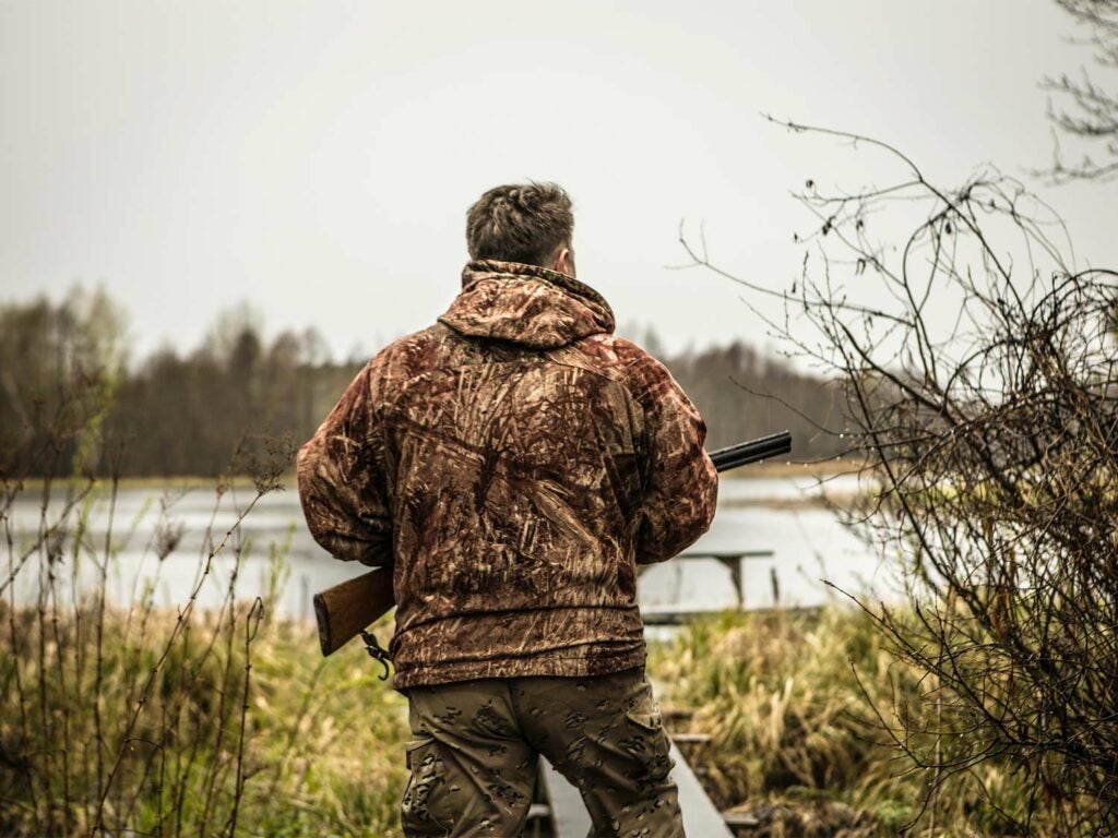 A hunter in camoflauge holds a shotgun while standing near a lake.