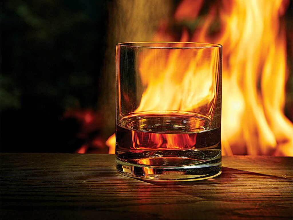 A glass of whiskey at night next to a campfire.
