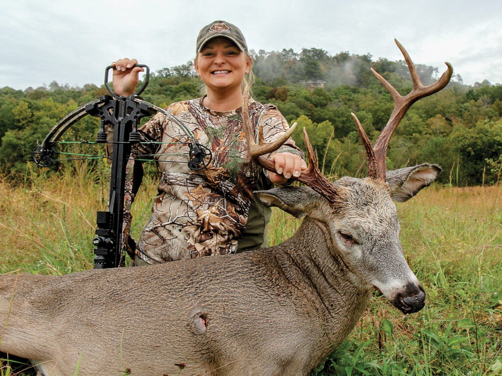 A woman hunter in full camo holds a crossbow while kneeling behind a large buck.