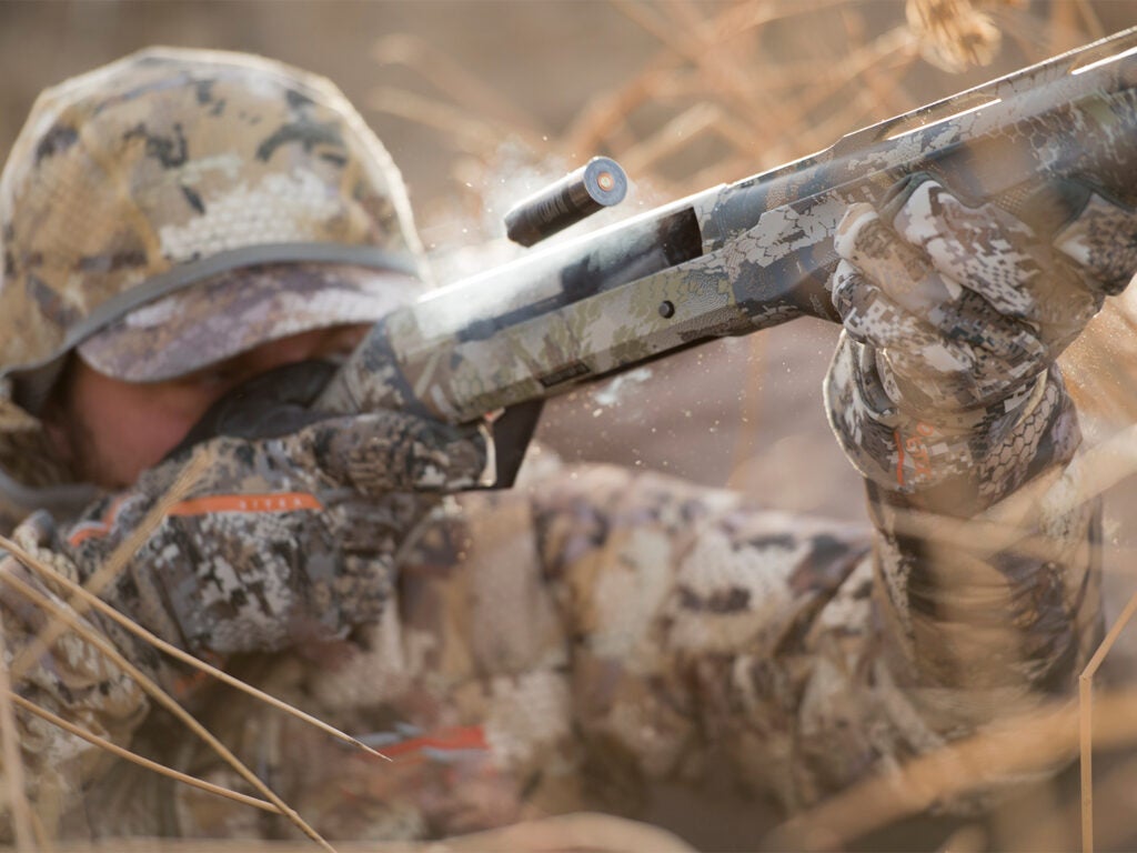 A hunter in full camo fires a shotgun in heavy brush.