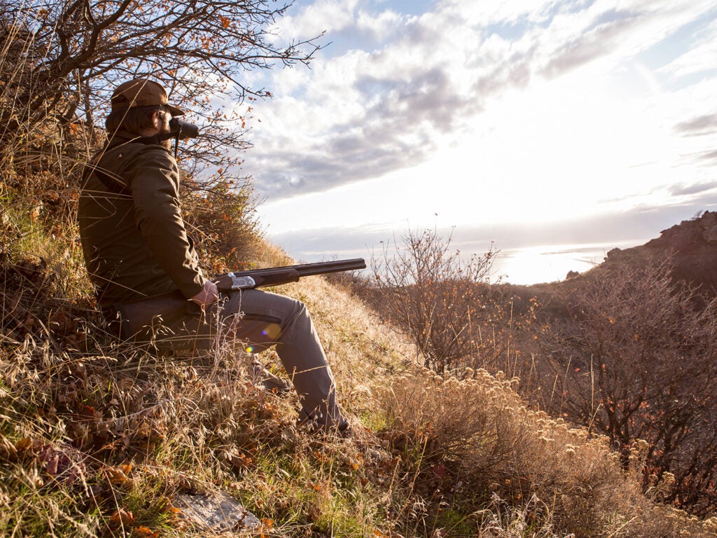A hunter sits on a hillside with a shotgun in his laugh while he's glancing through binoculars.