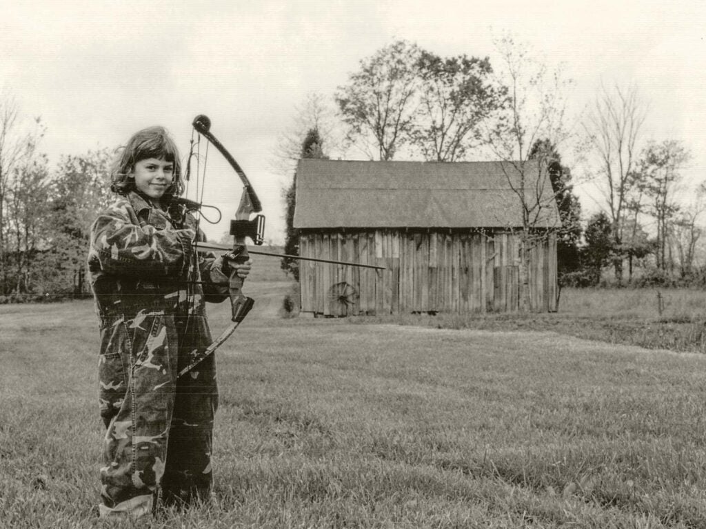 A black and white photograph of a young girl in oversized camoflauge coveralls while holding a large compound bow in front of old farm buildings.