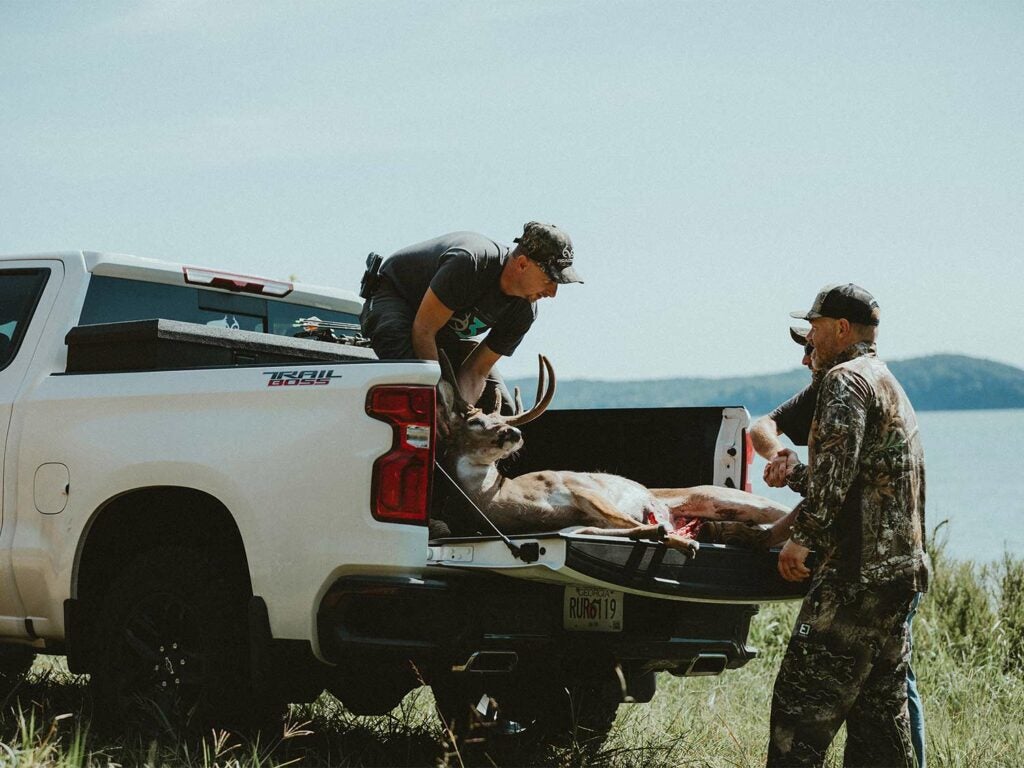 Two hunters pull a large whitetail buck into the bed of a truck.