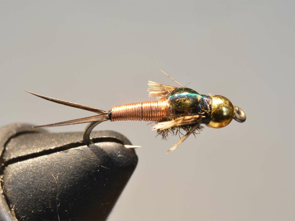 A copper, gold and green fly lure held in a vice.