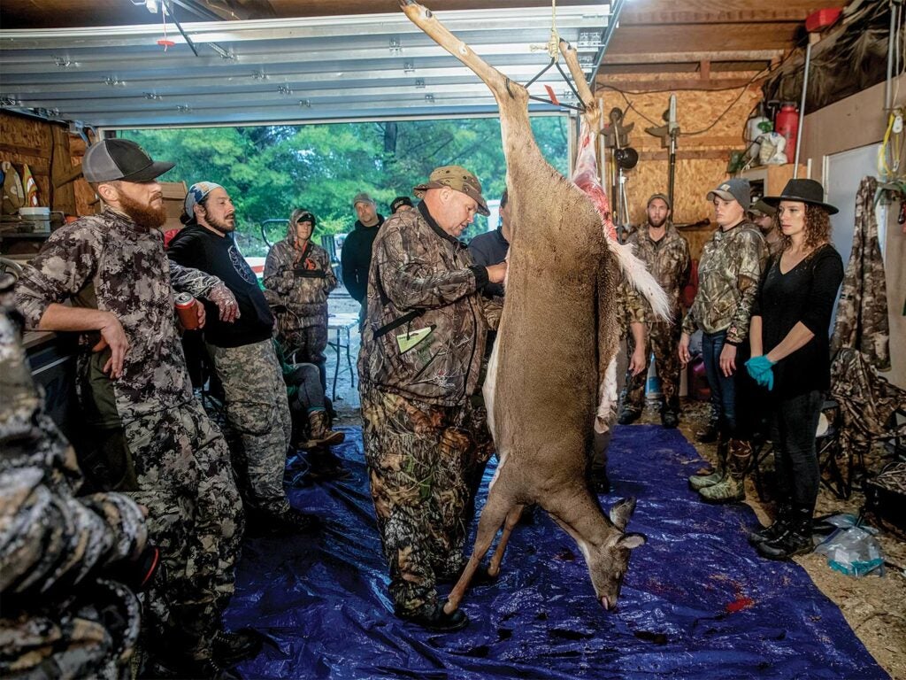 A group of hunters stand in a garage while one man butchers a deer hanging upside down on a y-hook.