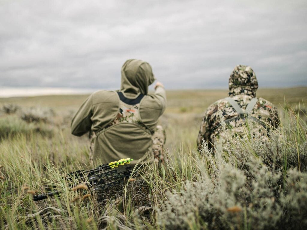 Two hunters sitting in tall grass in an open plain with gray clouds in the sky.