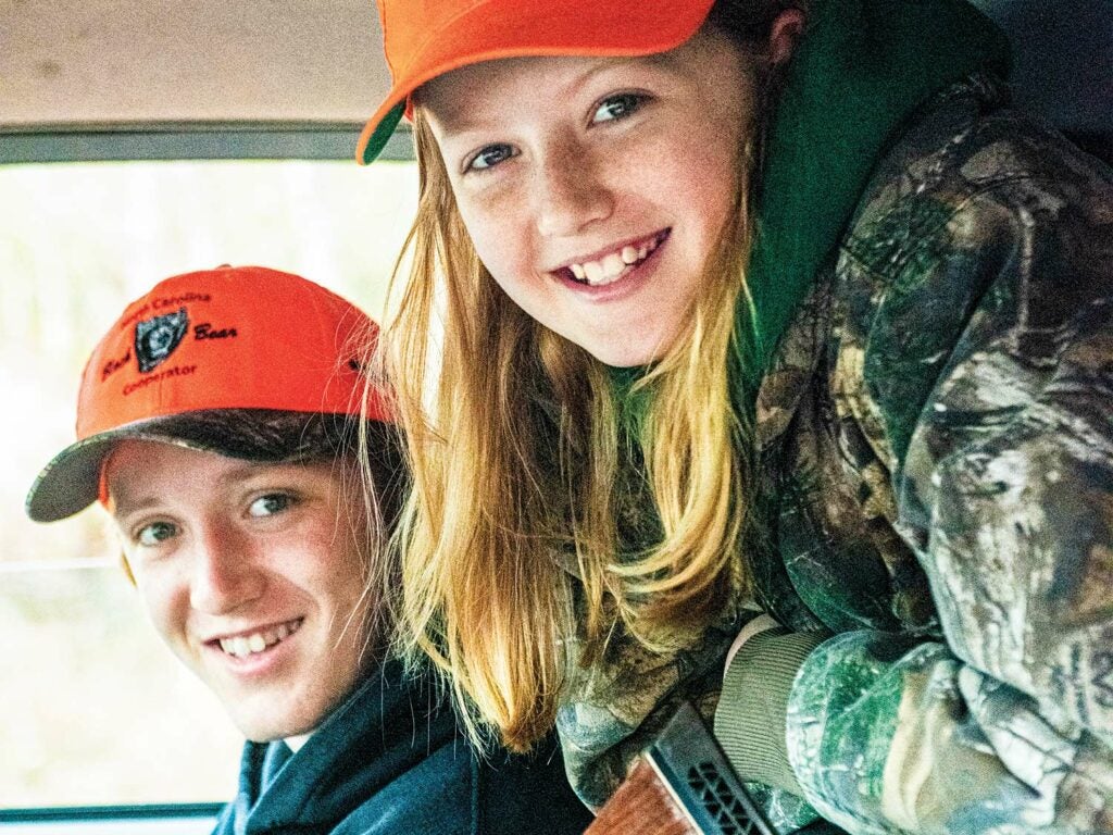 Two children, a bow and a girl, face the camera and smile while wearing hunting gear.