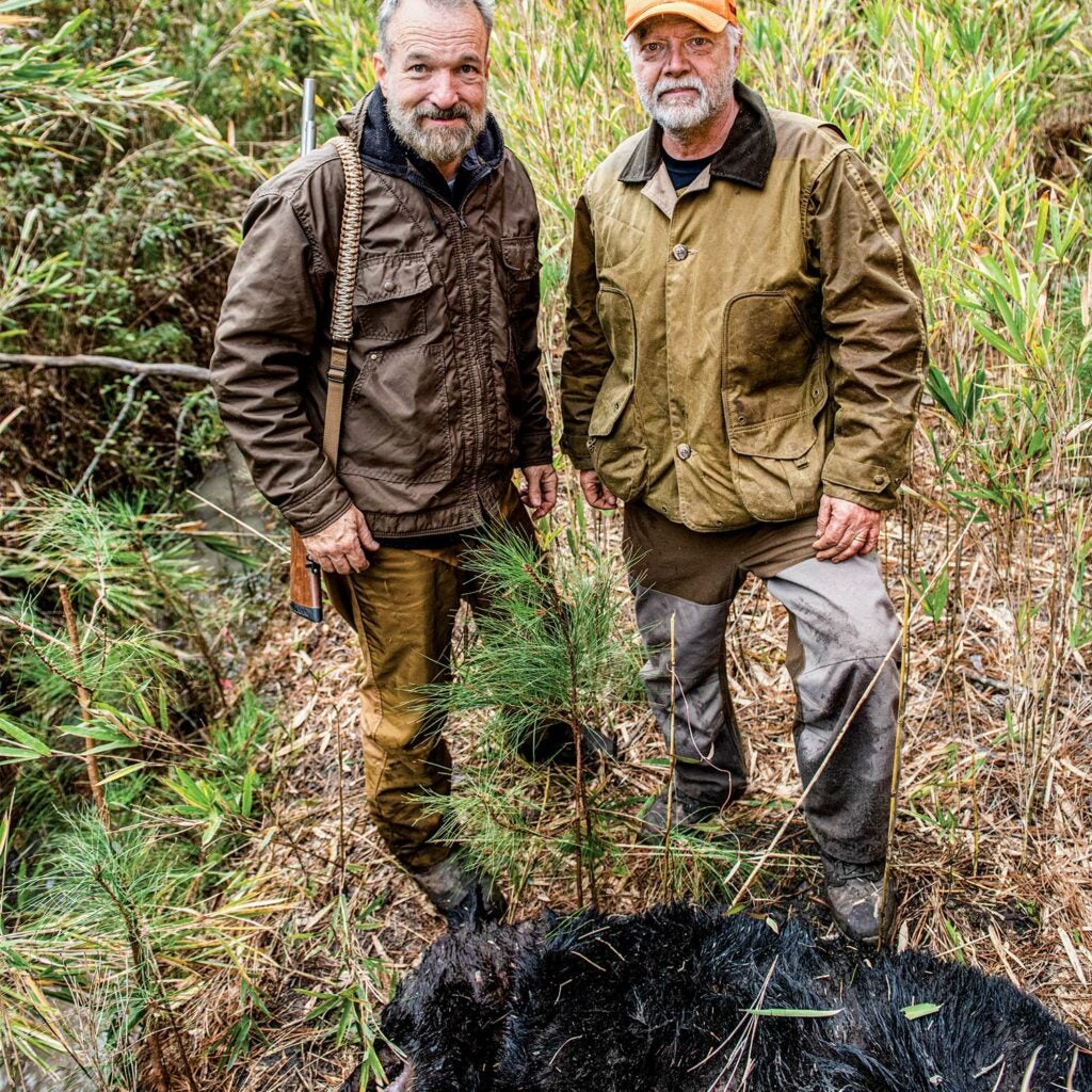 Two hunters stand behind a dead black bear and smile at the camera.