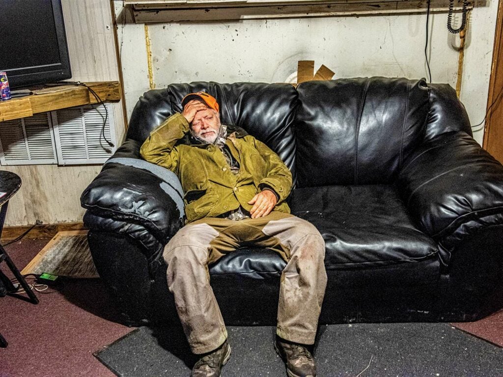 A tired male hunter relaxes on a black leather couch in a hunting cap.