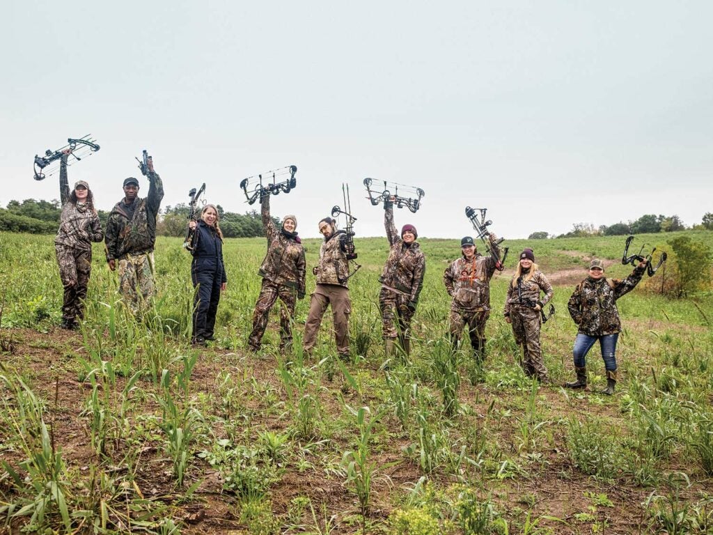 A group of nine hunters hold up their compound bows in celebration while standing in a large open field