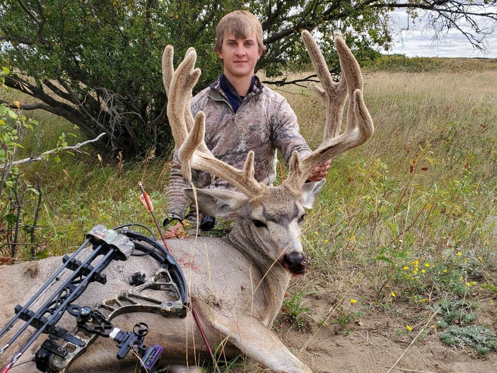 A young man kneels behind a mule deer and holds up its head by the antlers.