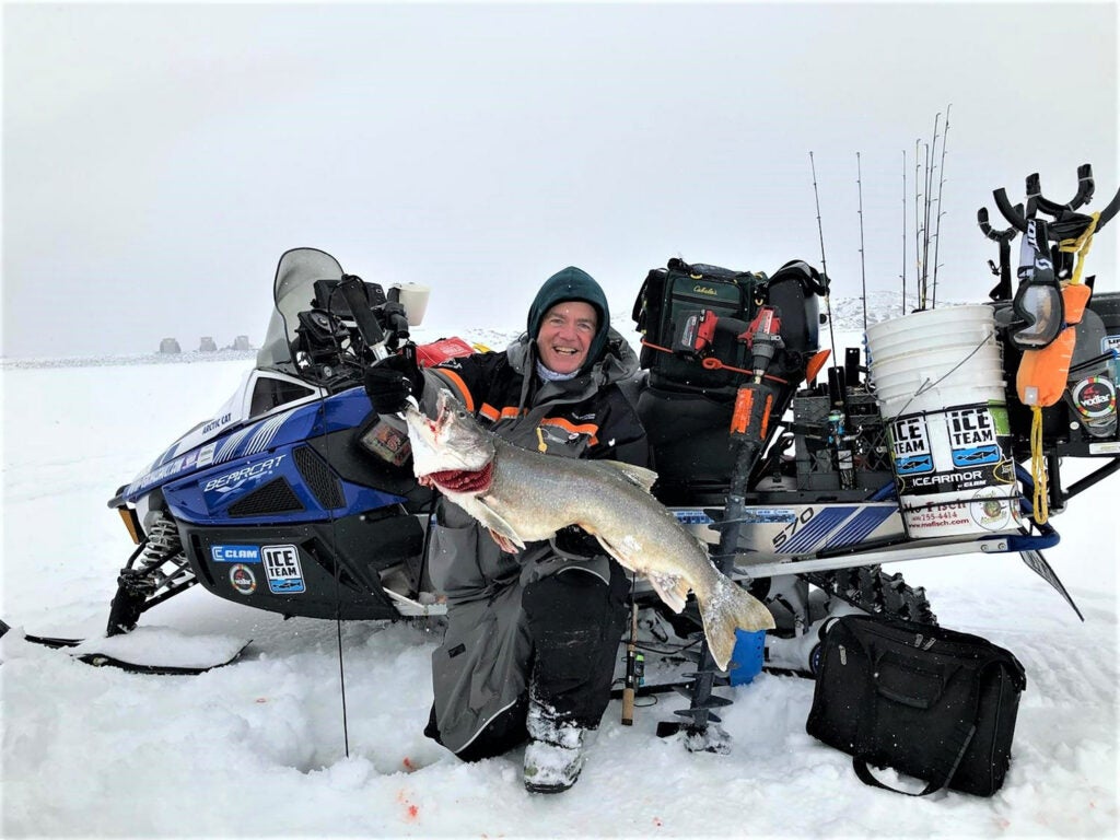 A man holds up a large fish pulled from ice fishing and is surrounded by fishing gear.