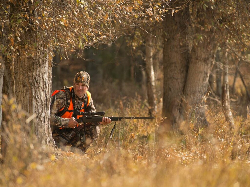 A hunter in full camo kneels behind a tree and grips a rifle in both hands.