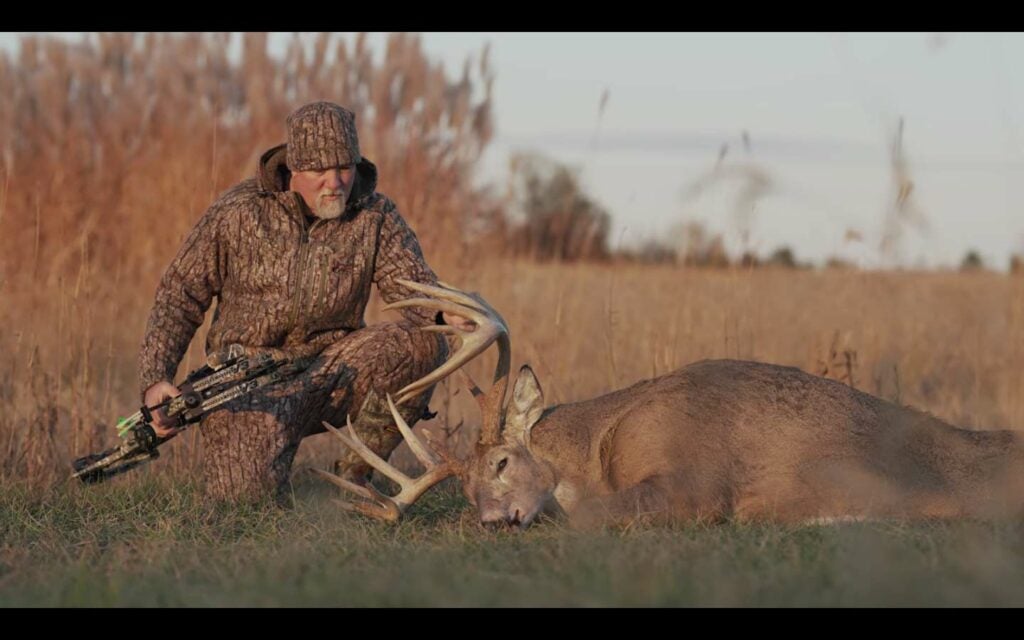 A hunter sits next to a dropped whitetail buck in a large open field.