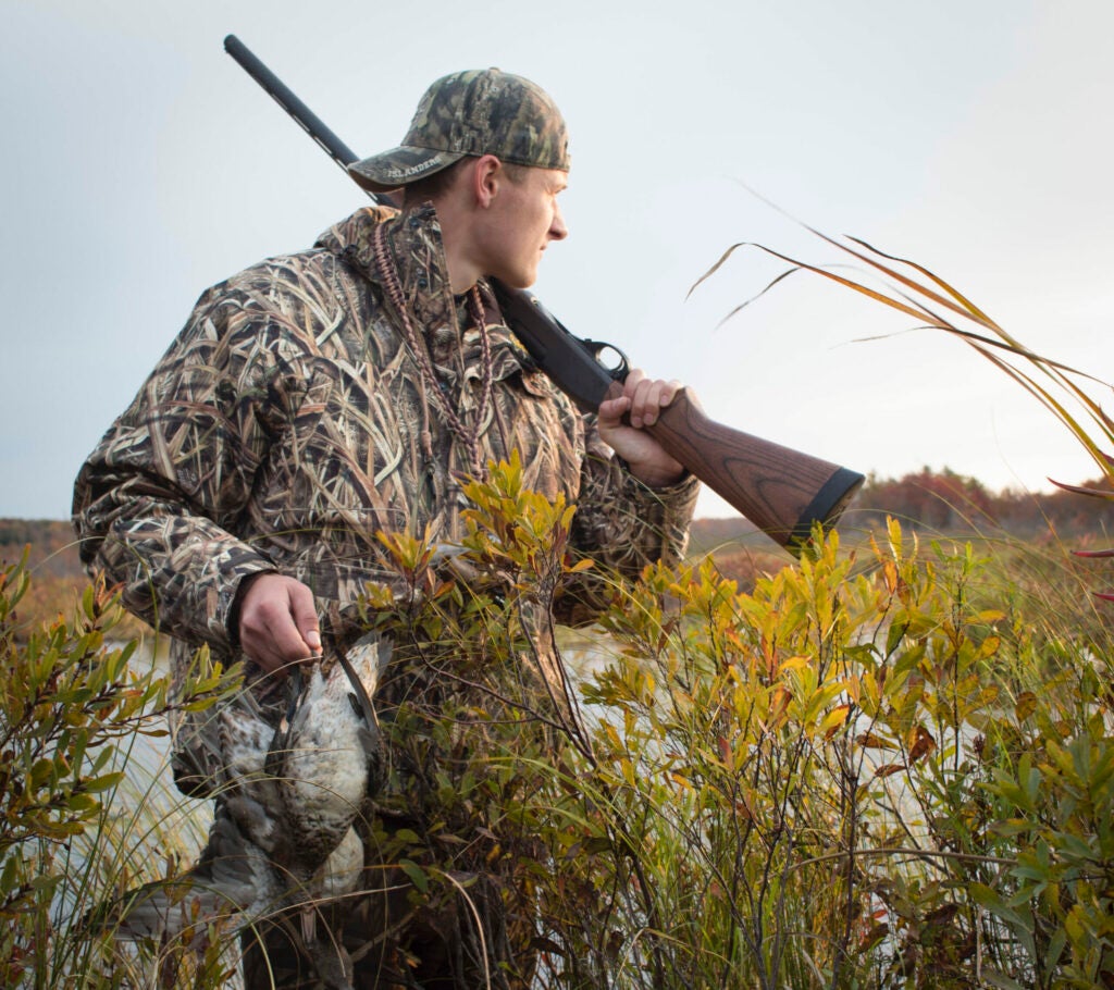 A hunter holds a rifle in one hand and a duck in the other.