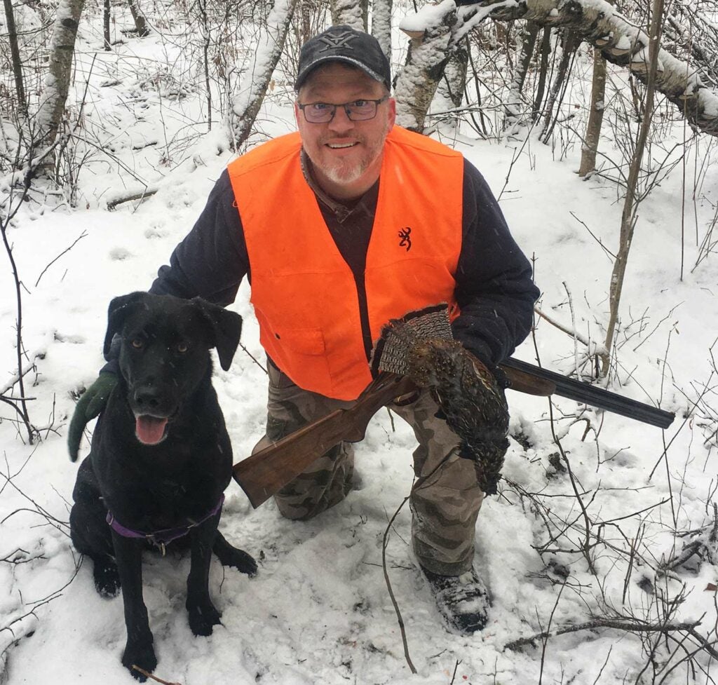 A hunter kneels in the snow and pets his hunting dog while holding a ruffed grouse.