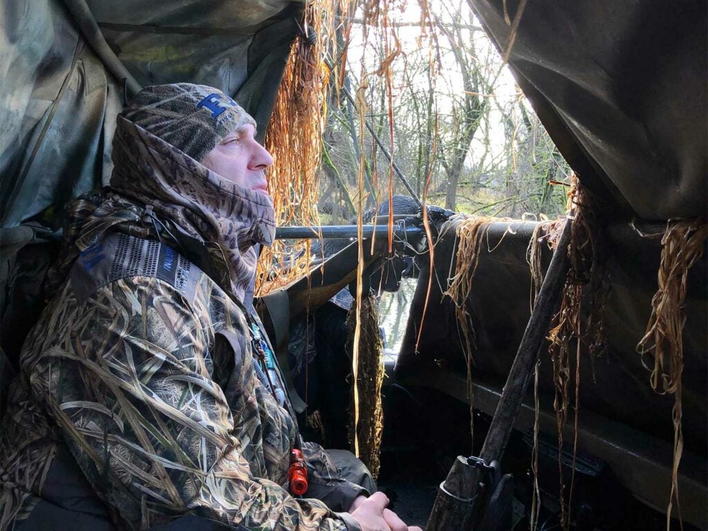 A hunter in full camo sits in a hunting blind while gazing out the window.