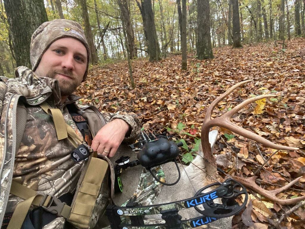 A hunter in the woods kneels next to a large buck in the woods.