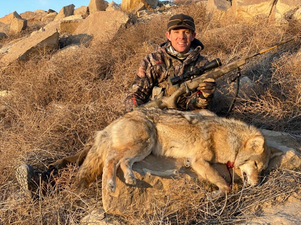 Hunter in rocky terrain holding a rifle and sitting behind a dead coyote.